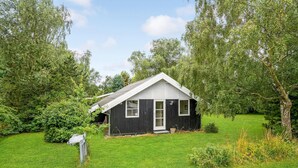 A small, single-story wooden cabin with a gabled roof, surrounded by lush greenery and trees.