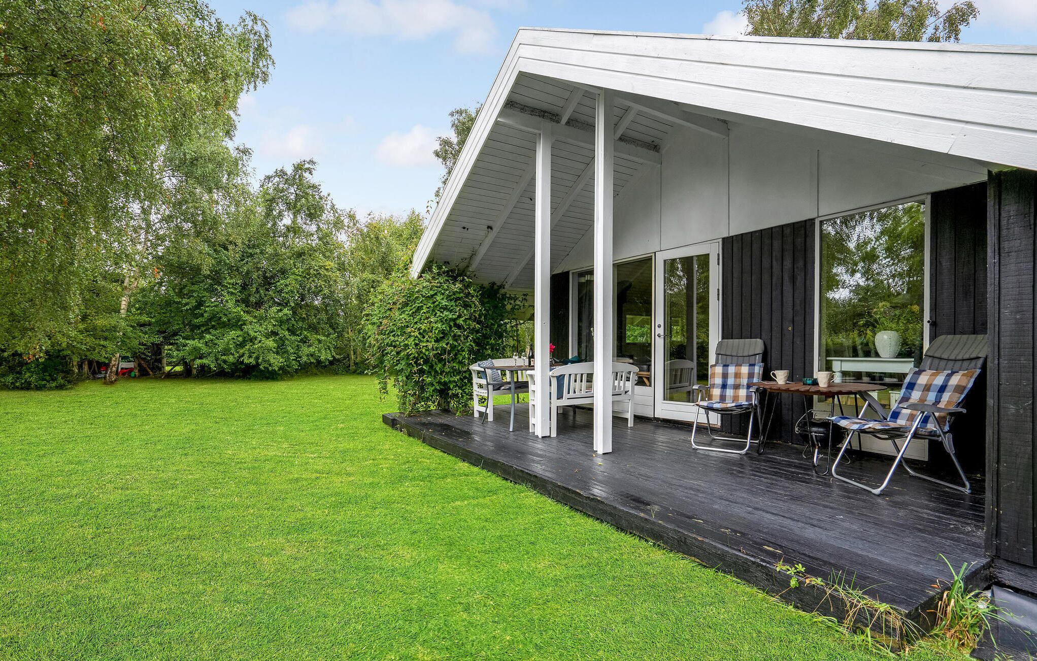 Un espace extérieur abrité doté d’une terrasse en bois, de mobilier blanc et offrant une vue sur une pelouse verdoyante et des arbres.