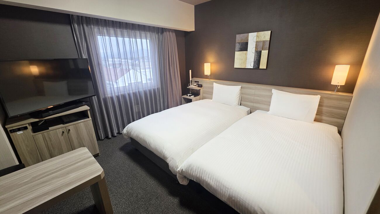 Quarto de hotel com cama, televisão, escrivaninha e janela com cortinas.