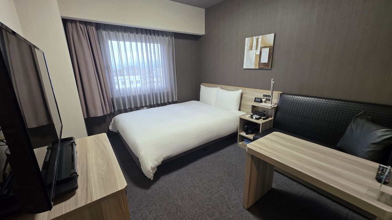 Quarto de hotel com cama, mesa de cabeceira, escrivaninha e janela com cortinas.
