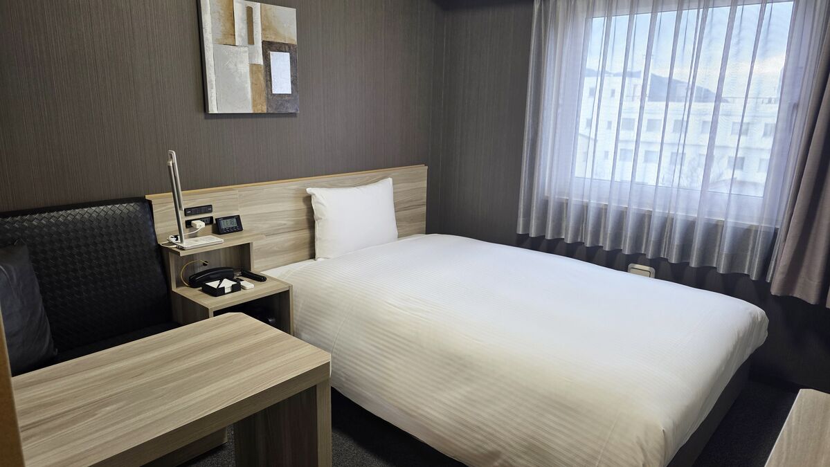 Quarto de hotel com cama, escrivaninha, cadeira, mesa de cabeceira com abajur, espelho e janela com cortinas.
