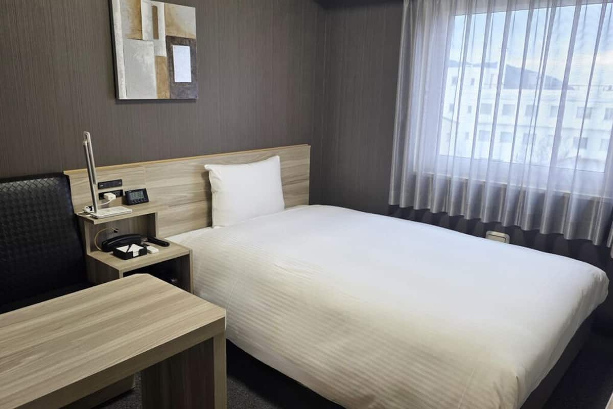 Quarto de hotel com cama, escrivaninha, cadeira, mesa de cabeceira com abajur, espelho e janela com cortinas.