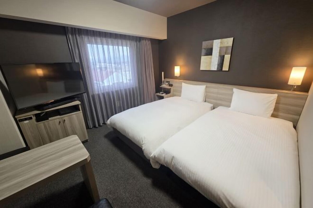 A hotel room with two beds, a TV, a desk, and a window with curtains.