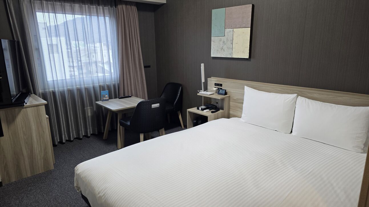 Quarto de hotel com cama, escrivaninha, cadeira, televisão e mesa de cabeceira com abajur.