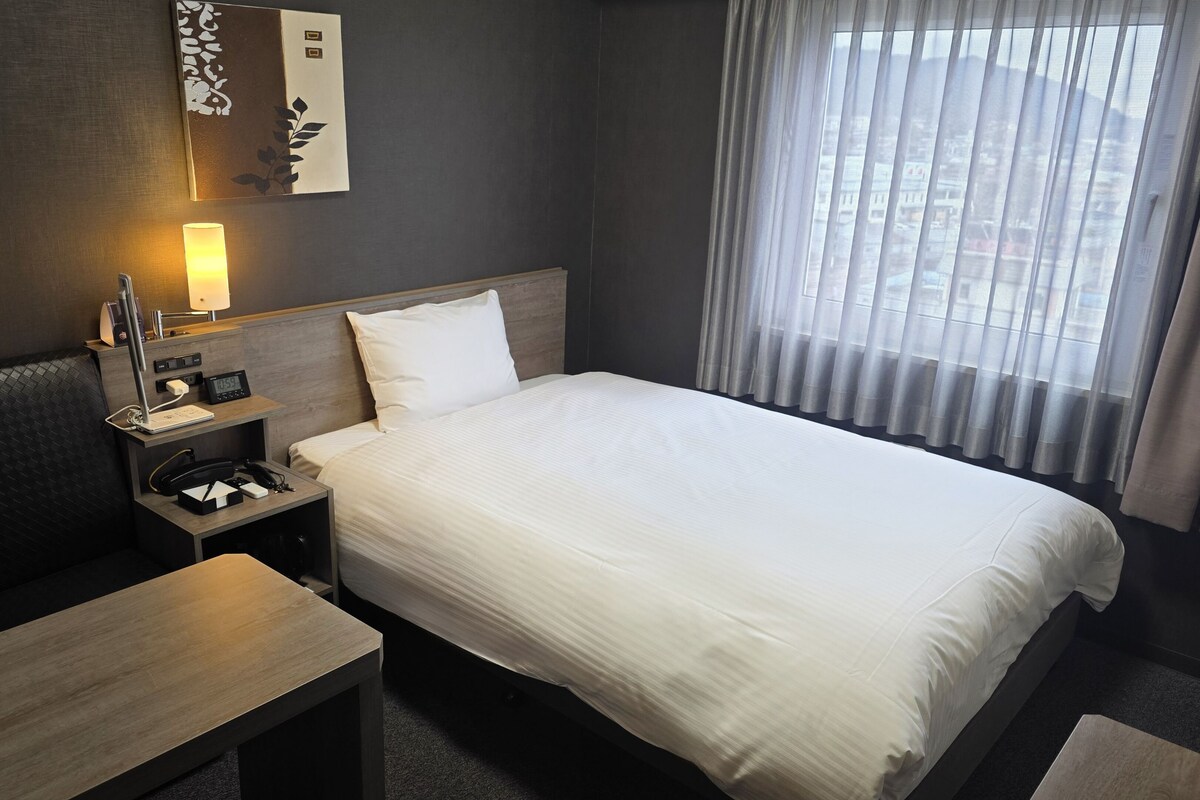 A hotel room with a bed, bedside table, lamp, and a window with curtains.