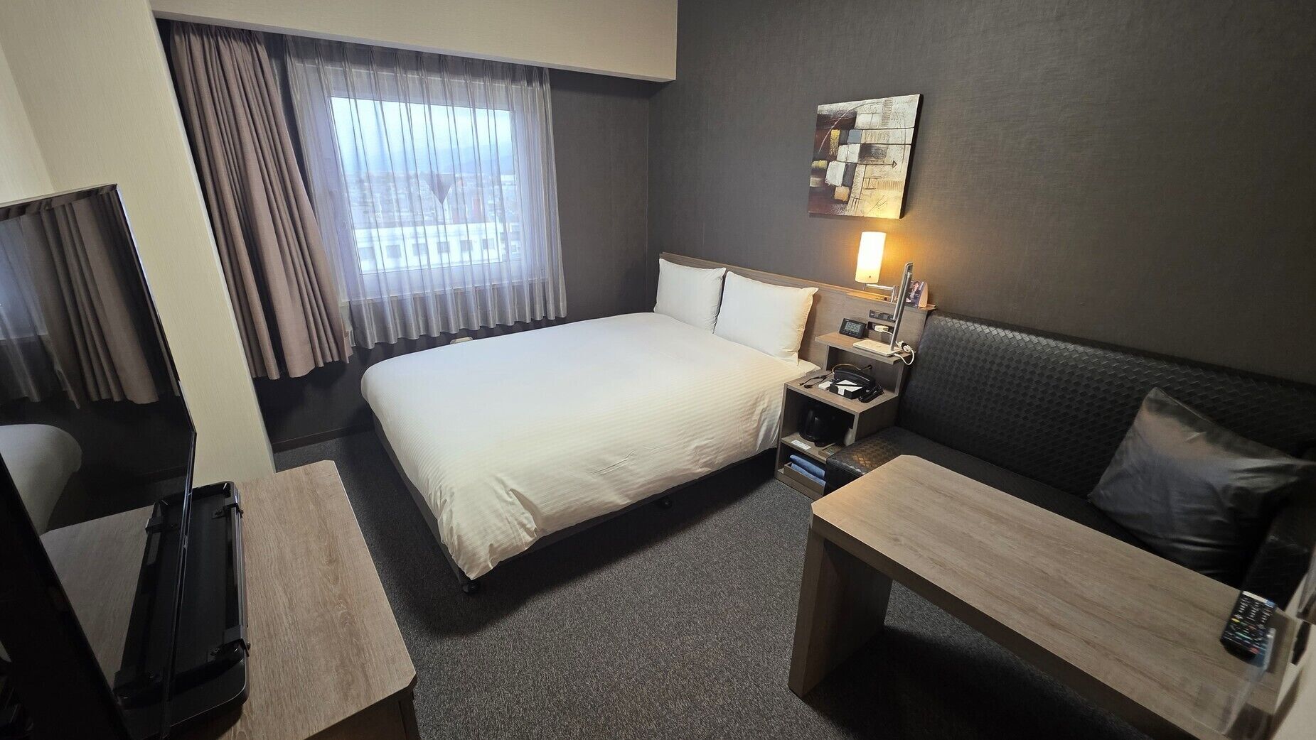 Quarto de hotel com cama, escrivaninha, sofá, mesa de cabeceira, abajur e um quadro na parede.