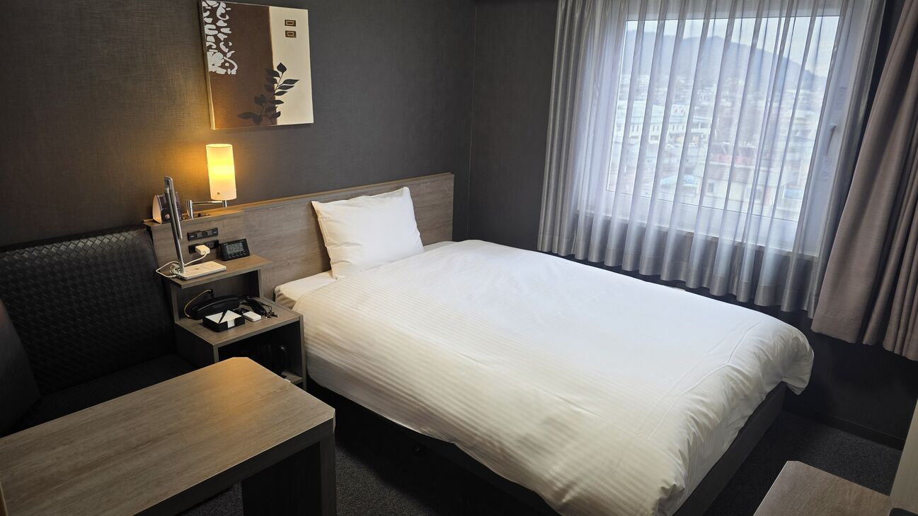 Quarto de hotel com cama, mesa de cabeceira, luminária e vista para a cidade pela janela.
