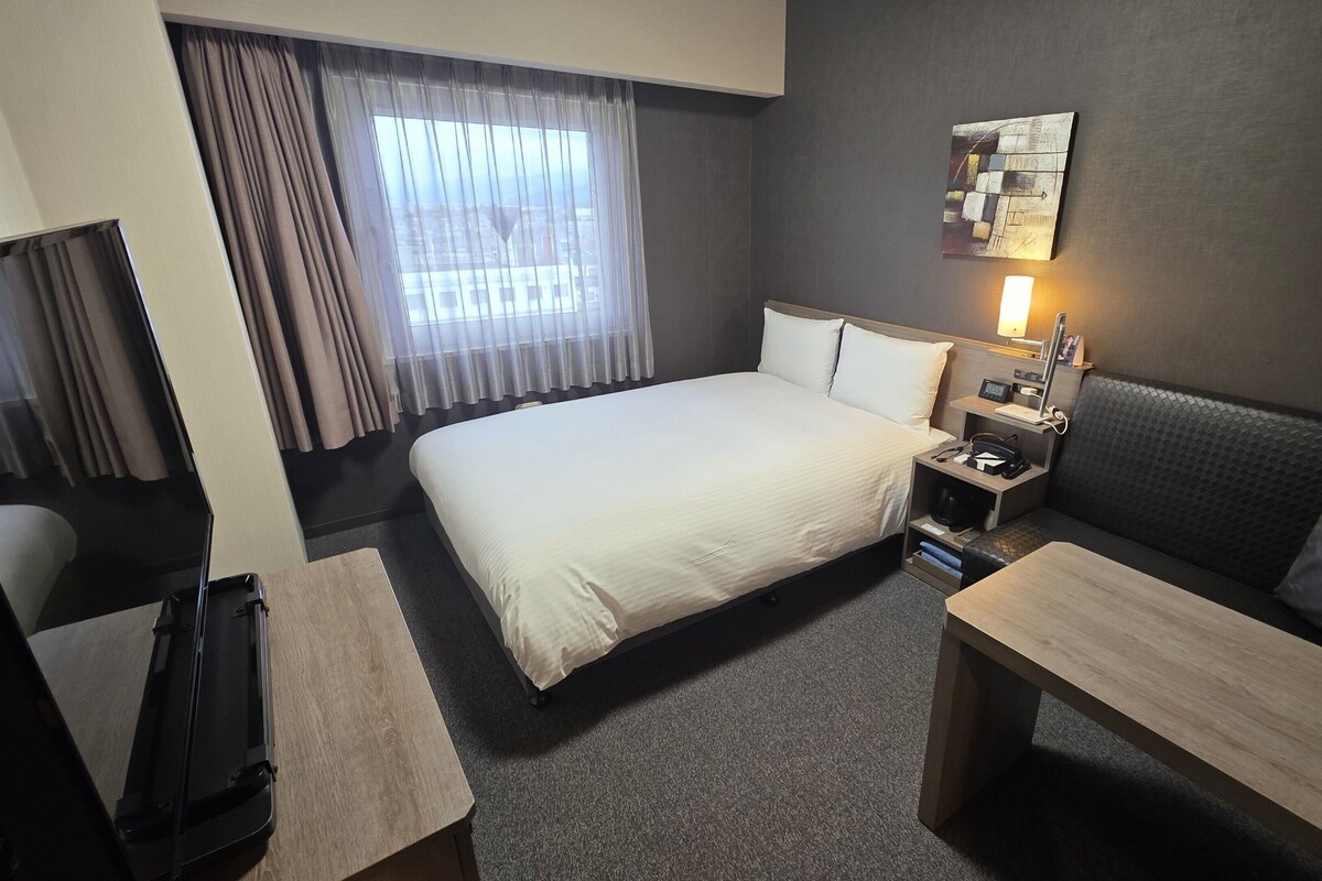 A hotel room with a bed, a desk, a sofa, a nightstand with a lamp, and a wall-mounted artwork.