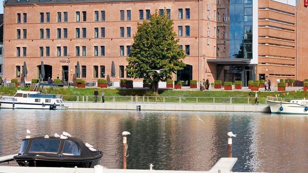 A hotel with a brick facade, a modern glass building to the right, and a tree in front. - Hotel Hafenspeicher - 14023 - Classic (Leer (Ostfriesland))