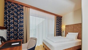 A hotel room with a bed, a desk with a chair, and a window with patterned curtains. - Hotel Hafenspeicher - 14023 - Classic (Leer (Ostfriesland))