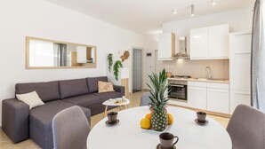 Ein modernes Wohnzimmer mit einem grauen Sofa, einem runden Esstisch mit Früchten und Tassen sowie einer Küche mit weißen Schränken und einem Herd.