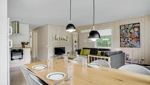 A modern kitchen with wooden paneling, a dining area with a wooden table set for four, and a living area with a sofa and a colorful abstract painting. - 3 bedroom amazing home in Hemmet (Hemmet)