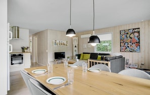 A modern kitchen with wooden paneling, a dining area with a wooden table set for four, and a living area with a sofa and a colorful abstract painting.