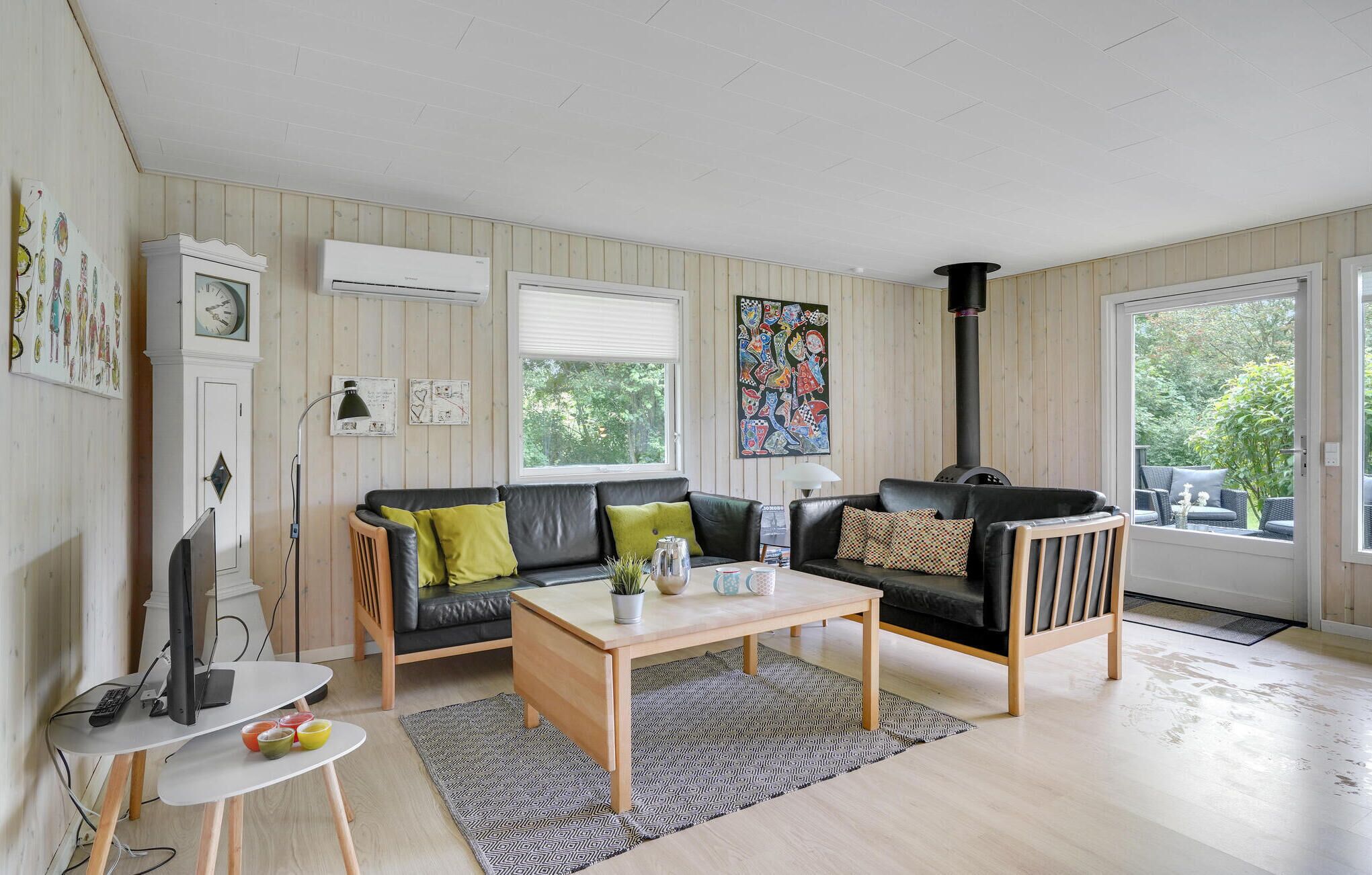 Een gezellige woonkamer met een zwarte leren bank, een houten salontafel en een zwarte fauteuil. Er hangt een witte wandklok, een schilderij en er is een raam met zonwering.
