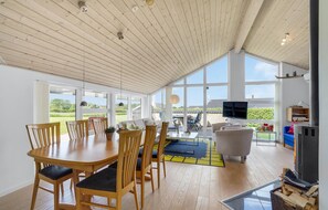 Una habitación amplia con techo de madera, ventanas grandes, un comedor con sillas de madera, un sofá y un televisor.