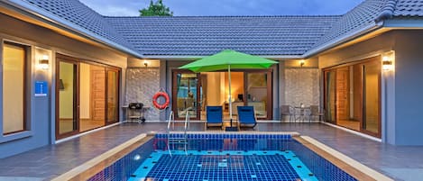 A swimming pool with blue tiles, surrounded by lounge chairs and a green umbrella.