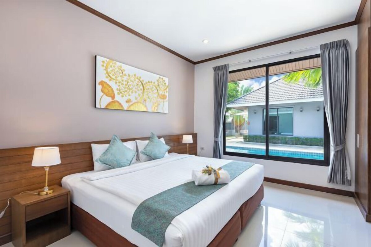 A hotel room with a large bed, a painting on the wall, a window with a view of a pool and a building, and a nightstand with a lamp.
