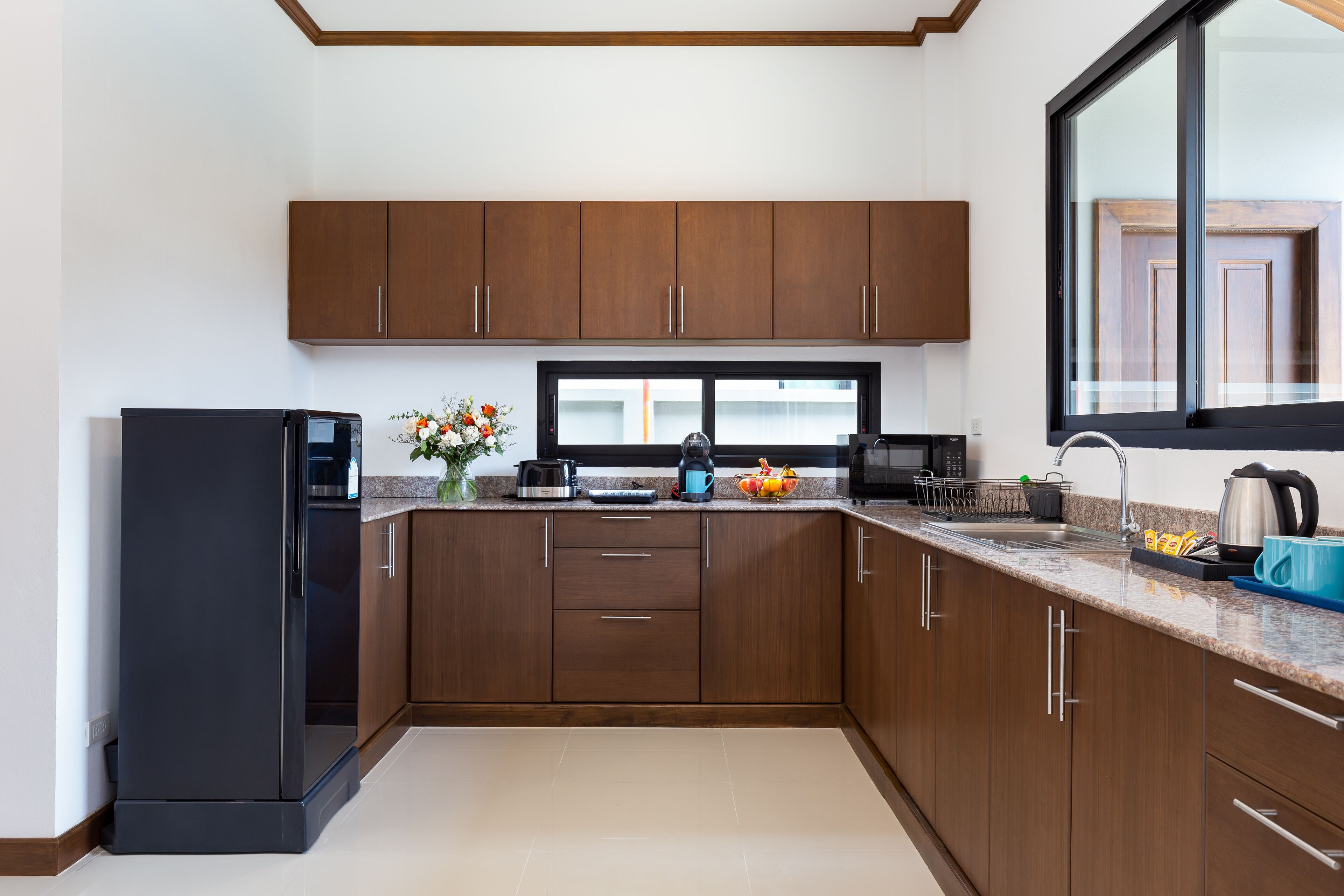 A modern kitchen with wooden cabinets, a black refrigerator, a window, and a countertop with various appliances and a vase of flowers.