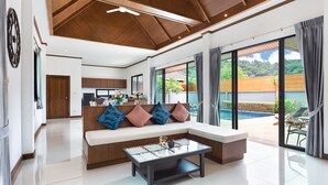 A modern living room with a large window, a sofa with cushions, a coffee table, and a view of a swimming pool. - Lady Naya Villas (Rawai)