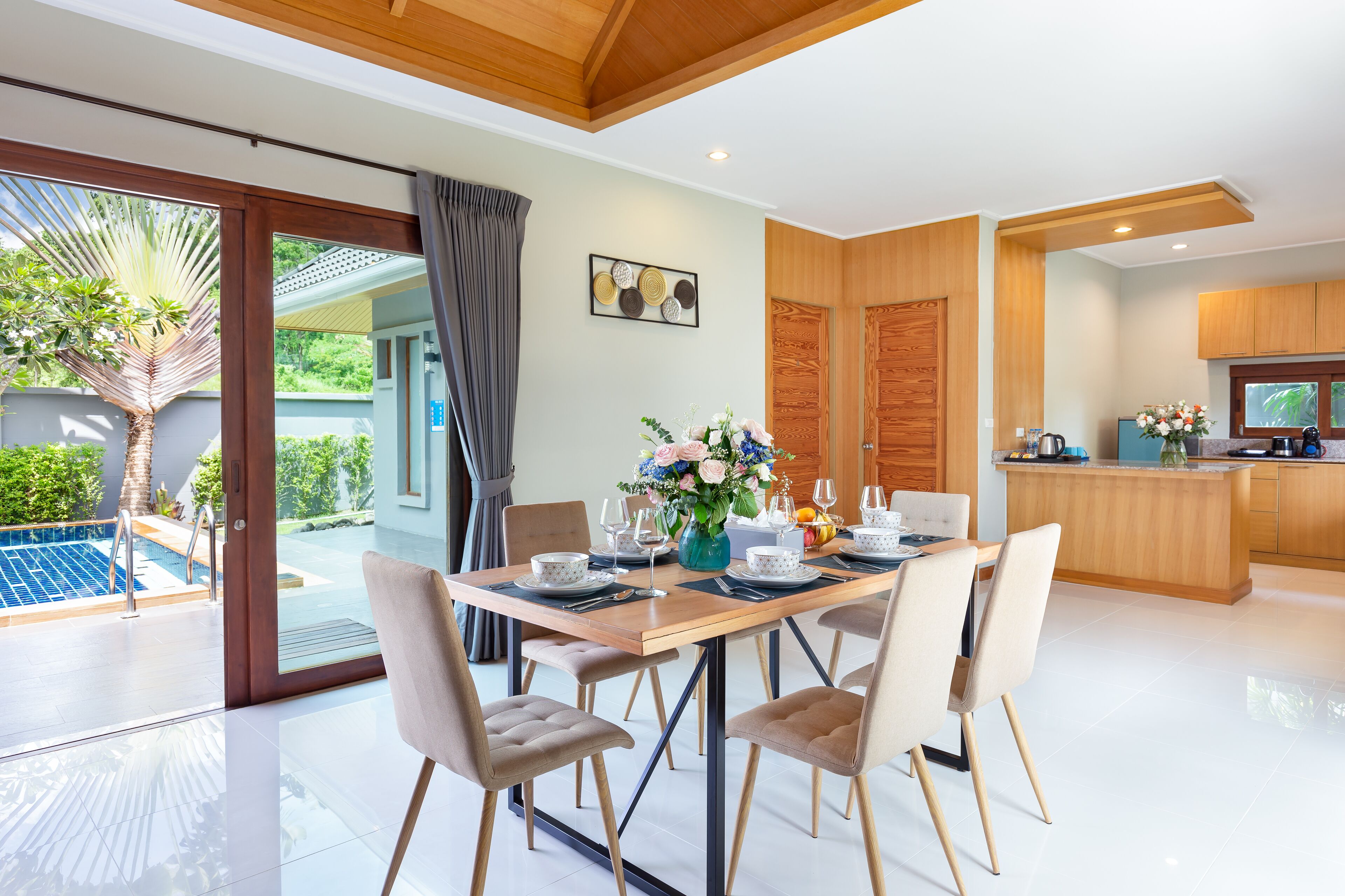 A modern kitchen with wooden cabinets, a dining area with a table set for four, and a view of a swimming pool outside.
