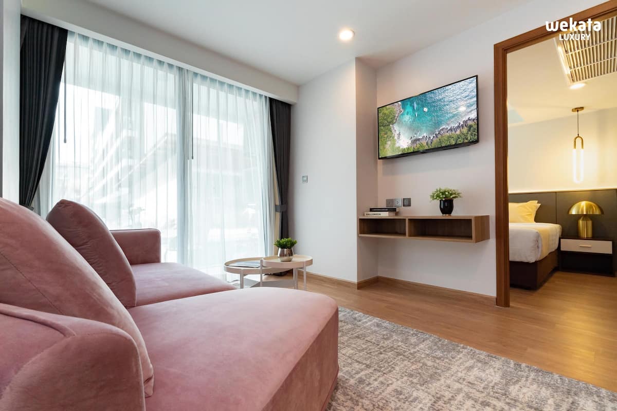Una sala de estar moderna con un sofá rosa, un televisor montado en la pared, una mesita con una planta y una cama con una mesita de noche.
