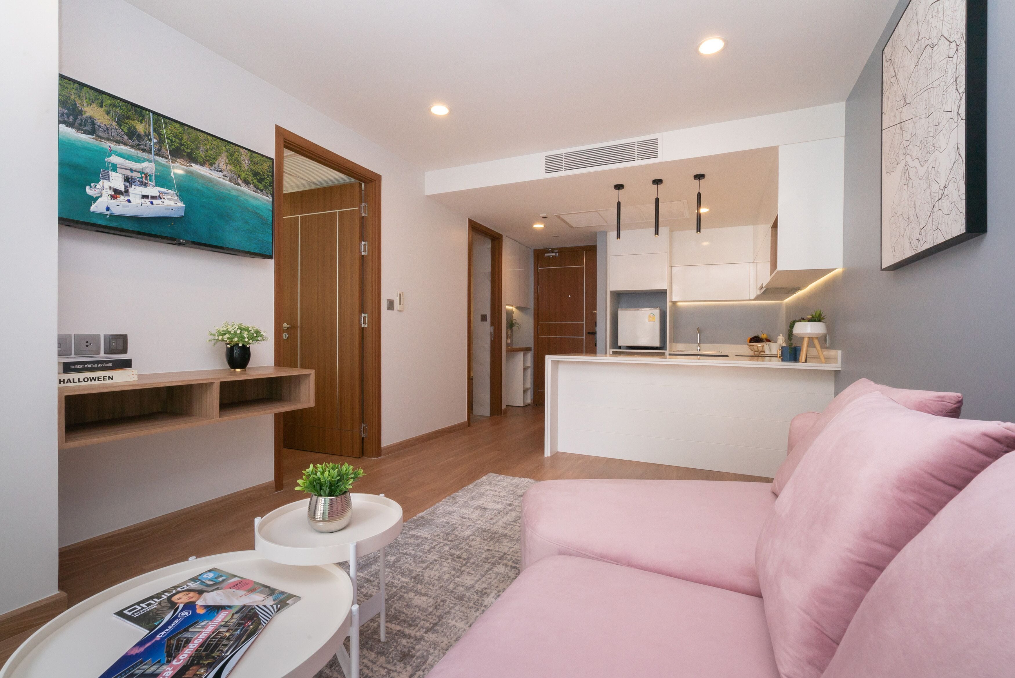 A modern living room with a pink sofa, a small white table with a magazine, and a potted plant. A kitchen with white cabinets and a microwave is visible in the background.