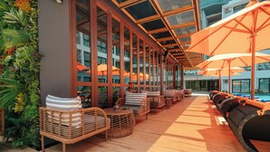 Un área de estar al aire libre con luz solar, mobiliario de mimbre y sombrillas naranjas, con vistas a una piscina.