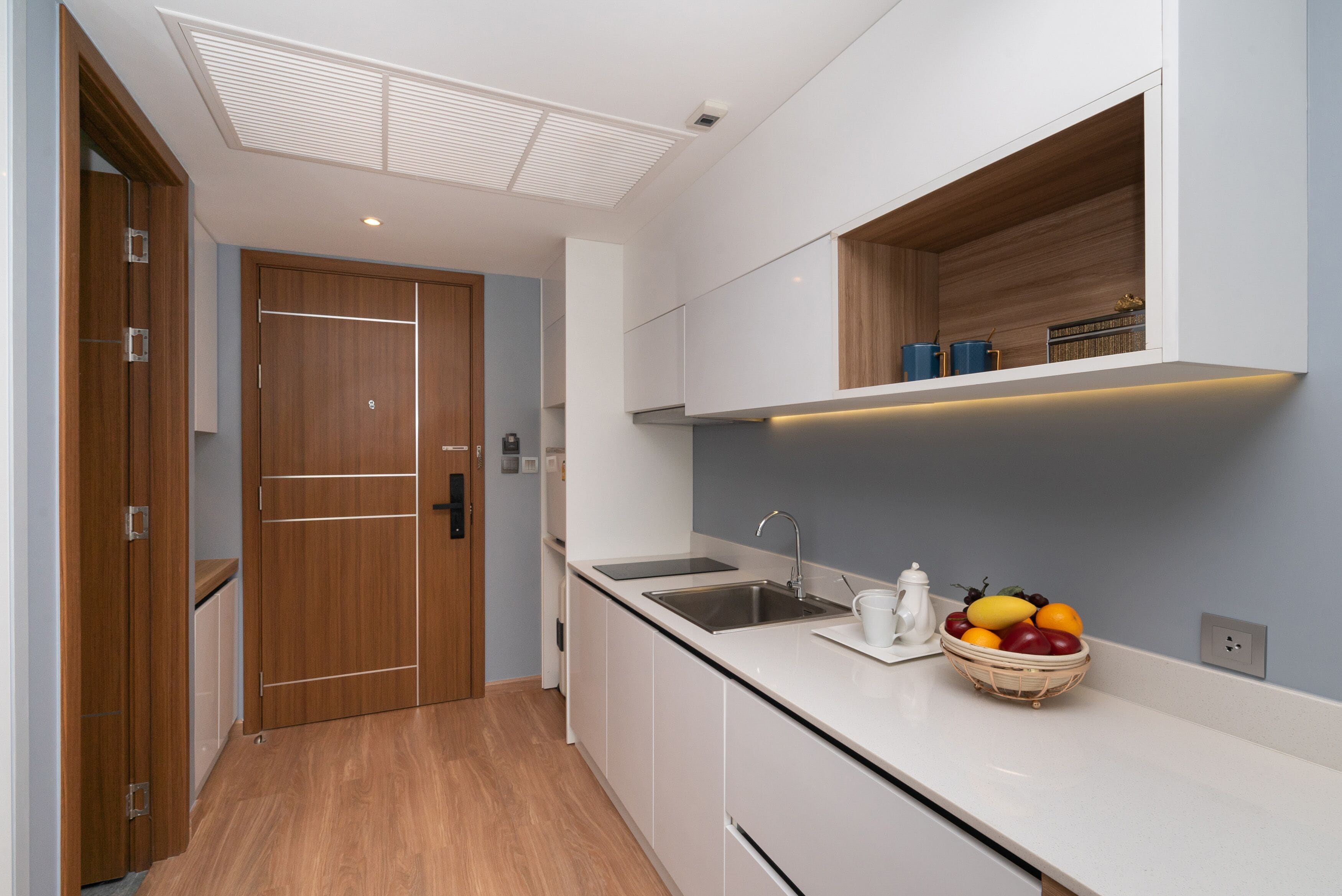 A modern kitchen with a wooden door, white cabinets, a built-in sink, and a fruit bowl on the counter.