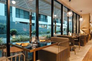 A modern restaurant with glass tables, comfortable seating, and large windows offering views of the building and greenery outside.