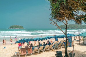 A beach with rows of sun loungers and umbrellas, a small island in the background, and the ocean waves. - Wekata Luxury (Karon)