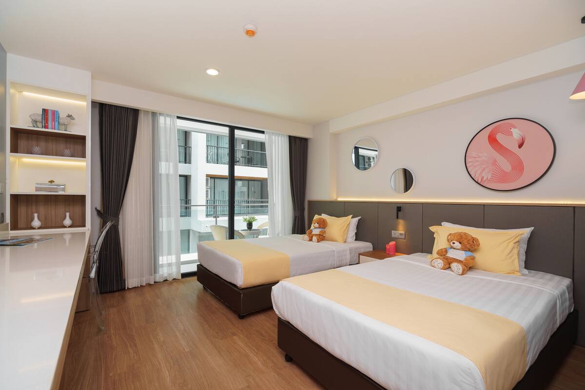 Habitación de hotel con dos camas, un escritorio, una estantería y un ventanal con cortinas.
