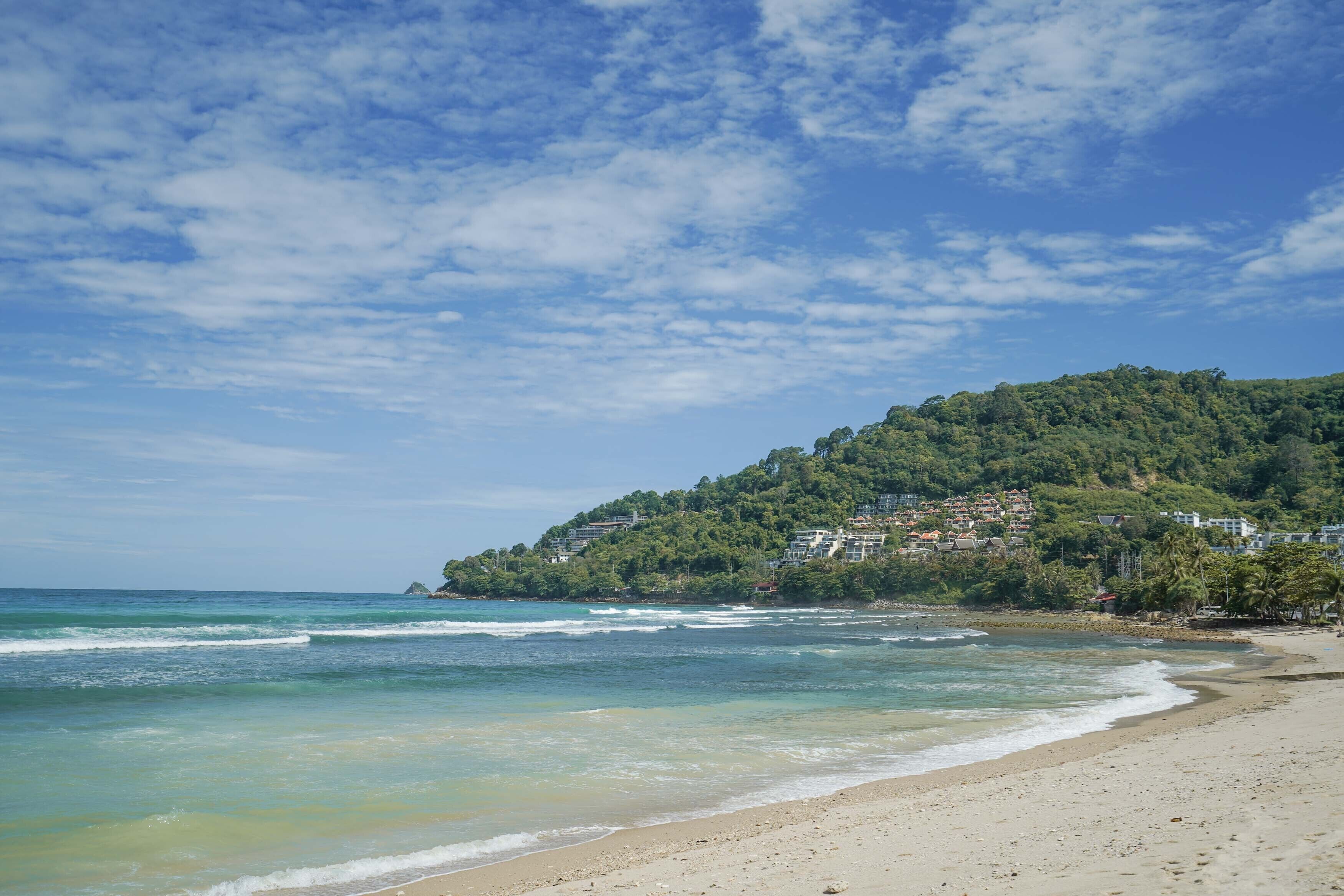 A beach with clear waters, a sandy shore, and a hill with buildings and trees.