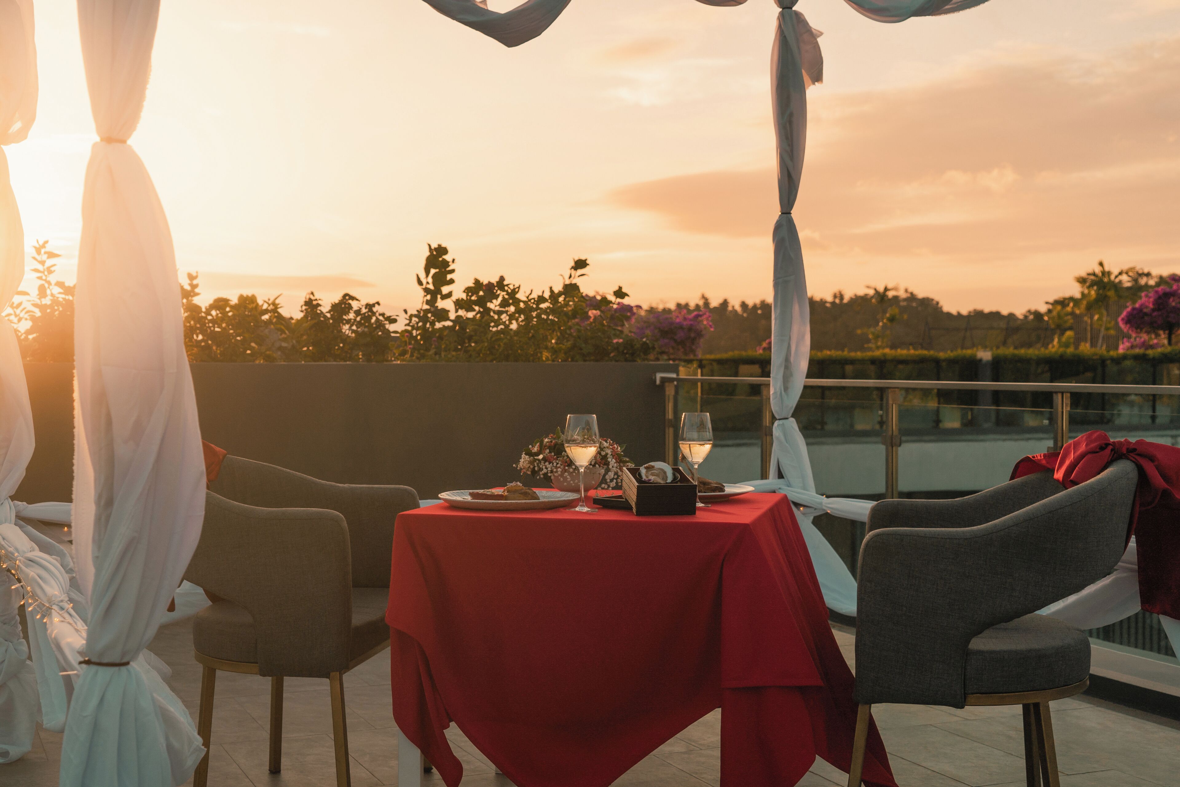 Mesa posta para dois com toalha vermelha, duas cadeiras e vista para o pôr do sol sobre a água.
