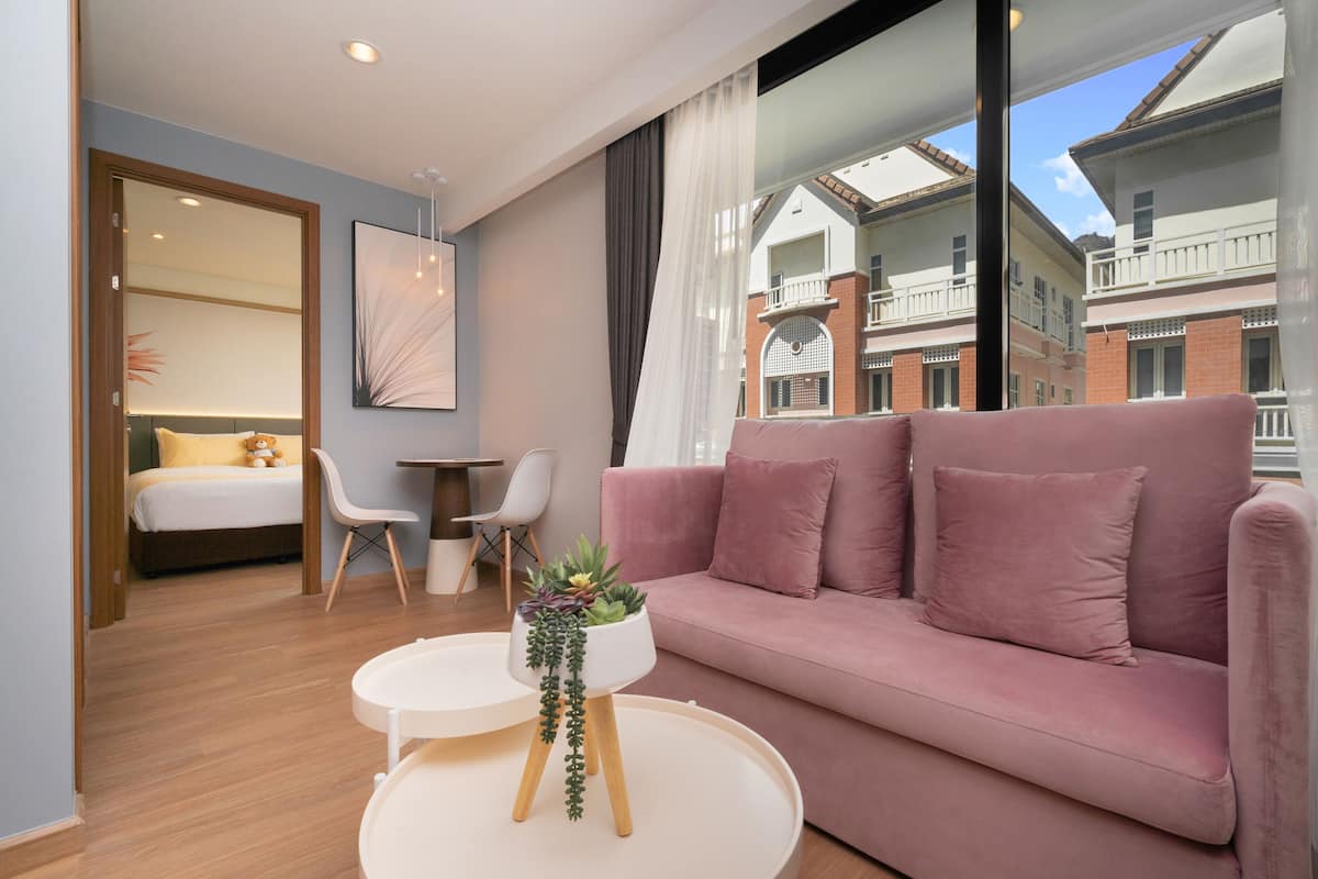 Una sala moderna con un sofá rosa, una mesa de centro redonda con una planta, una cama con cabecera blanca y vistas a edificios a través de ventanas amplias.