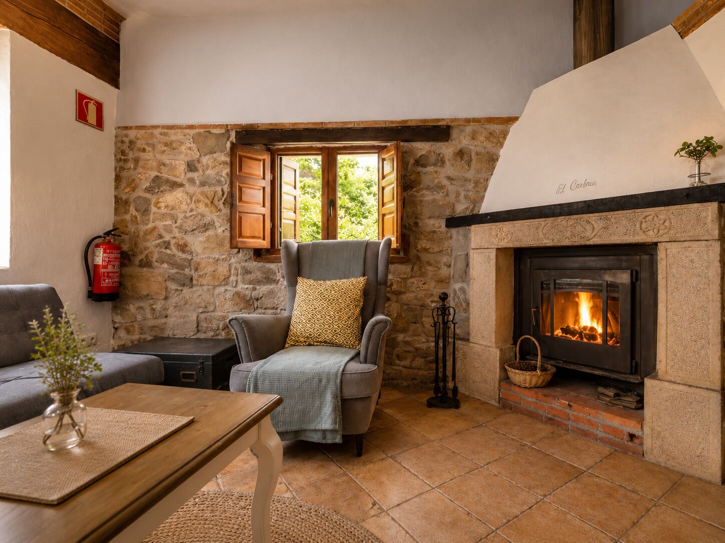 A cozy room with a stone wall, a fireplace, a comfortable armchair, a small table, and a window with shutters.