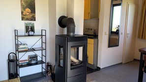 A compact living space with a wood-burning stove, a bookshelf with books, a kitchen area, and a door leading to another room.