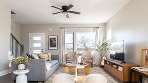 Un salon avec un canapé gris, deux fauteuils, une table basse, un téléviseur et un ventilateur de plafond.