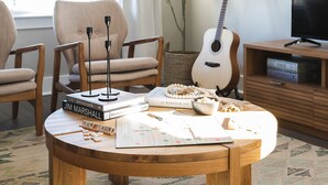 Un salon chaleureux avec une table basse ronde en bois, deux fauteuils, une bibliothèque remplie de jeux de société et une guitare.