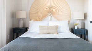 Un lit bien fait, avec du linge de lit blanc et un plaid gris, flanqué de deux tables de chevet ornées de lampes et d’objets décoratifs.