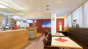 Un restaurante moderno con mostrador de recepción de madera, zona de estar con mesas y sillas, un cuadro grande en la pared y un reloj.