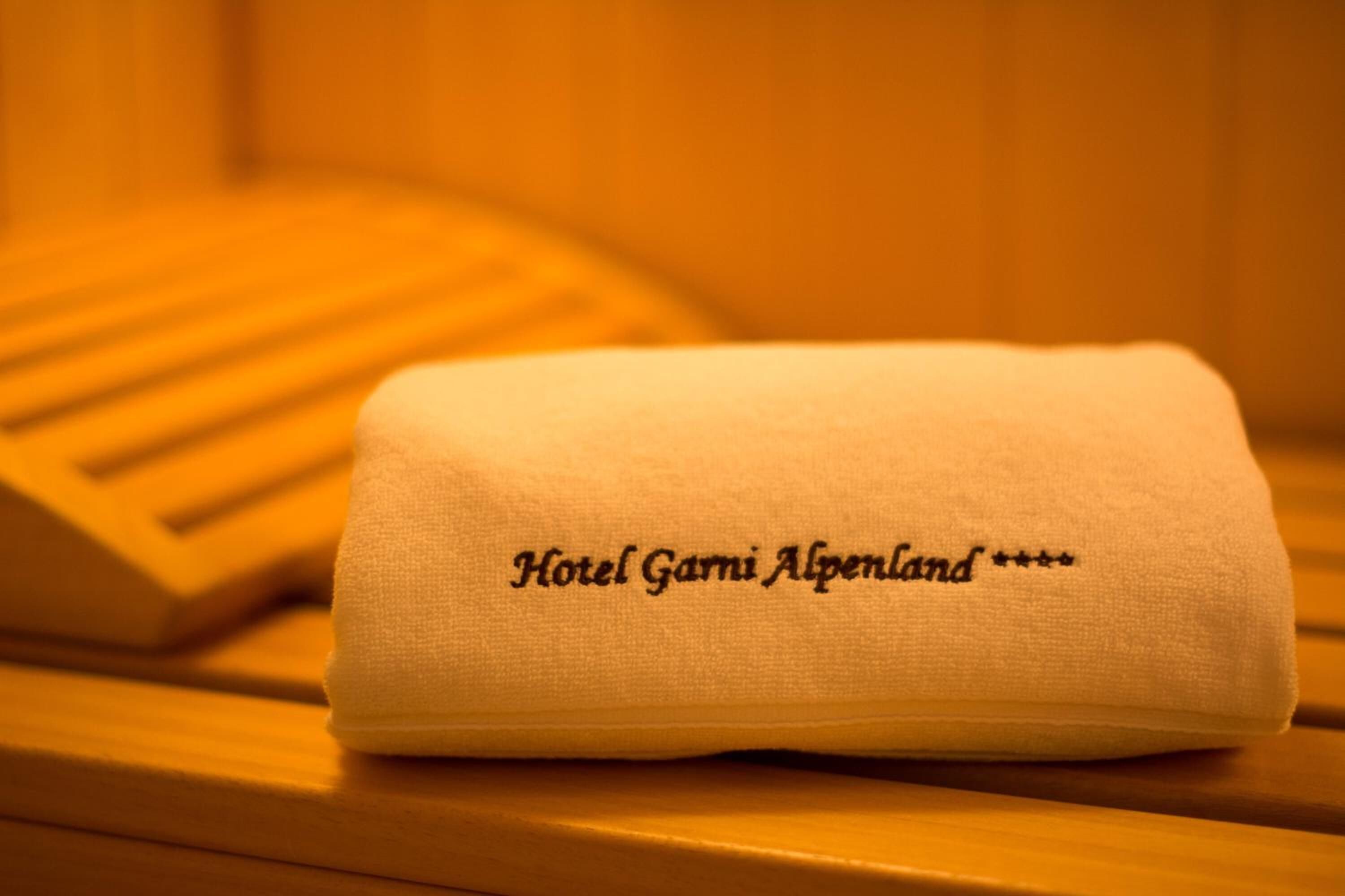 Une serviette blanche pliée, ornée d’une broderie représentant le nom « Hotel Garmi Alpenland », est posée sur une surface en bois.