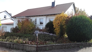 Una casa con tetto in tegole, circondata da un giardino con diverse piante e un vialetto lastricato.