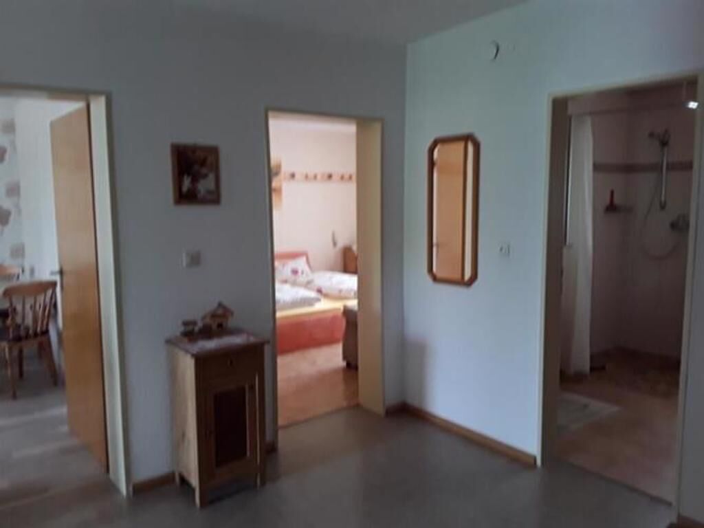 A room with a bed, a small table, and a mirror on the wall. There is a door leading to another room with a shower.