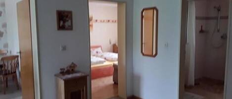 A room with a bed, a small table, and a mirror on the wall. There is a door leading to another room with a shower.