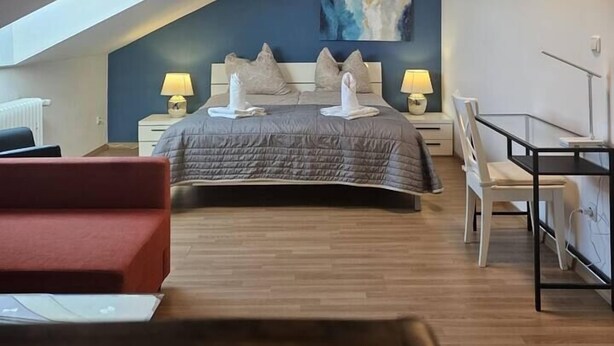 Un dormitorio con techo inclinado, una cama con almohadas, dos mesitas de noche con lámparas, un escritorio y una silla roja.