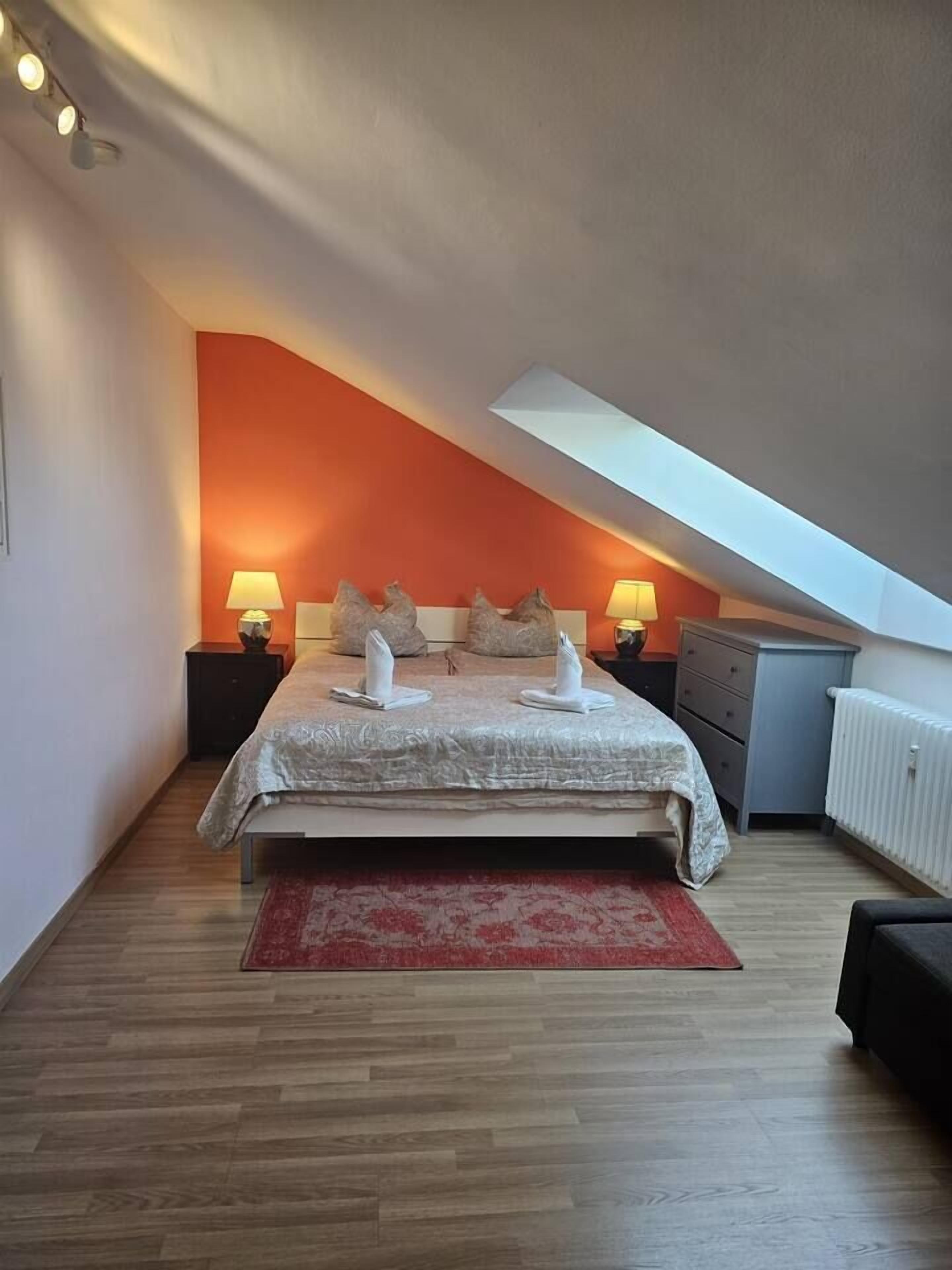 Een slaapkamer met een schuine kap, een bed met een witte dekbedovertrek, twee nachtlampjes en een rood tapijt.