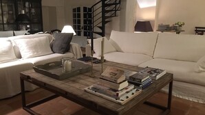 A cozy living room with a white sofa, a wooden coffee table with books and a candle, and a staircase in the background. - Charming house on the Ile de Ré (Loix)