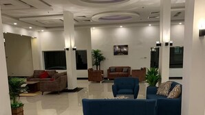 Ein geräumiger Eingangsbereich mit einem blauen Sofa, einem braunen Sofa und einem blauen Sessel. Im Hintergrund befinden sich Kübelpflanzen und ein großes Fenster.