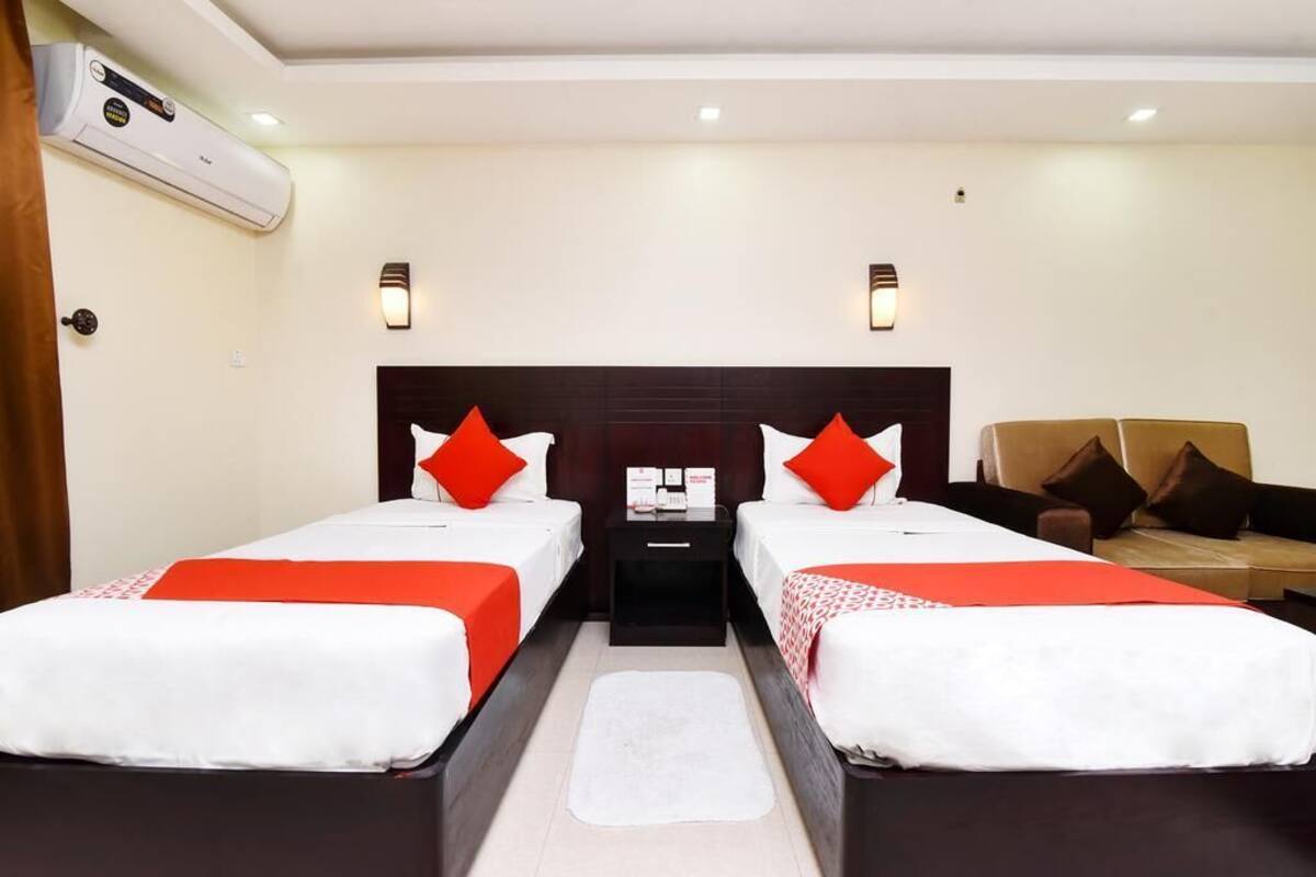 A hotel room with two single beds, each with red and white bedding, a nightstand with a lamp, and an air conditioning unit on the wall.
