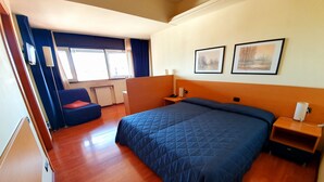 Quarto de hotel com uma cama grande, mesa de cabeceira com abajur, uma cadeira e janela com cortinas.